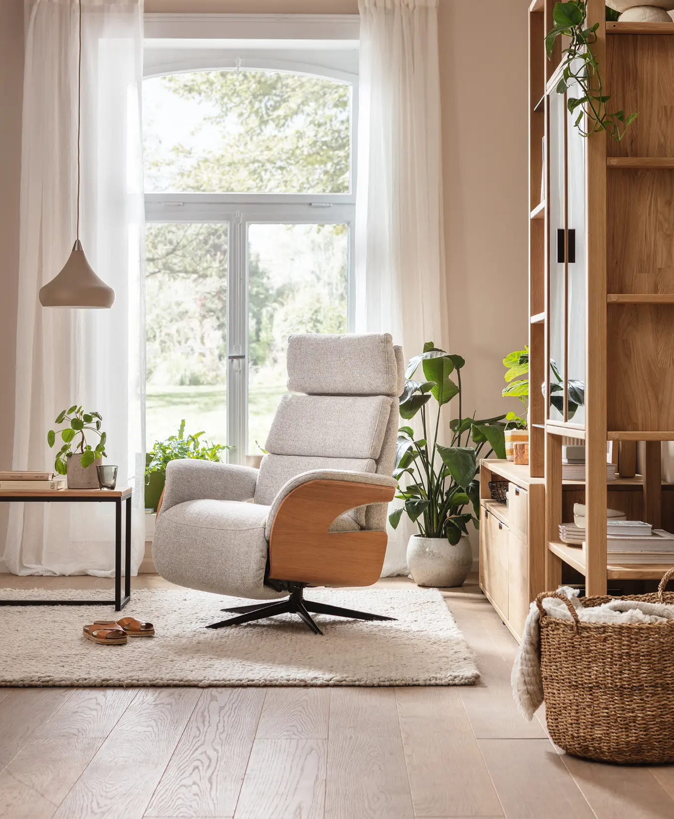 Polstersessel mit Holzarmlehnen steht vor großem Fenster mit Vorhängen, Pflanzen und Regal rechts im Wohnzimmer